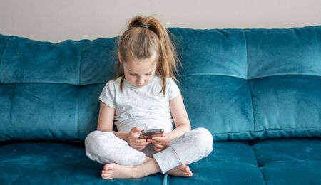 A little girl is watching cartoons on her phone while sitting on the sofa. children and gadgets.の写真素材