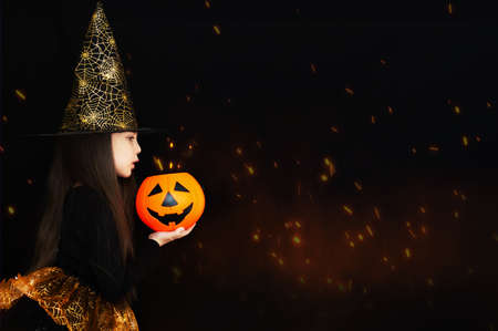 A little girl in a witch costume, holding a pumpkin, shot on a black background. Happy Halloween concept.の写真素材