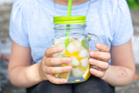 A small, beautiful white girl in a dress, in the summer, drinks cooling lemonade outdoors. Portrait of a smiling girl with her hair down.の写真素材