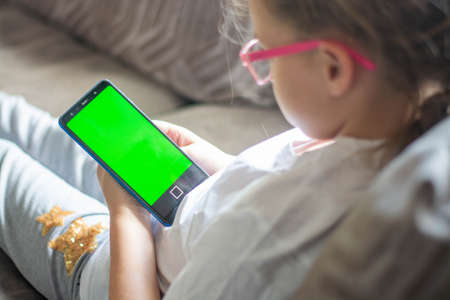 A happy little girl looks at the green screen of her mobile phone and laughs at a funny movie. a snapshot of a mobile phone monitorの写真素材