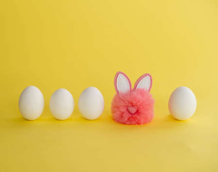 Minimalistic Easter composition. White eggs stand in a row, among them a pink fluffy ball with...