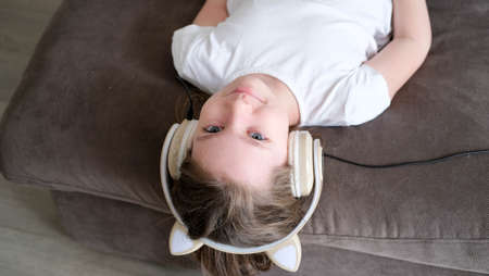 A small, white girl listens to music with headphones, lying on the sofa in the living roomの写真素材