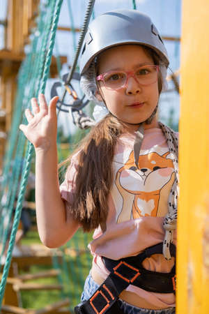 A brave young Caucasian girl in a helmet and insurance, climbing a rope town, in an amusement park on summer vacation, in a childrens camp. The concept of childrens recreation in summer.の写真素材