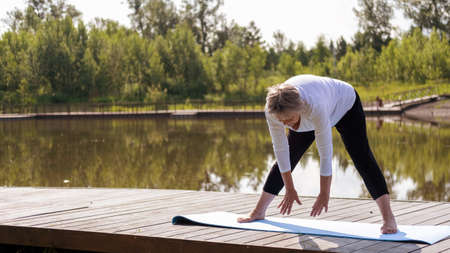An elderly Caucasian, gray-haired woman does stretching exercises in the park on a yoga mat. Outdoor...