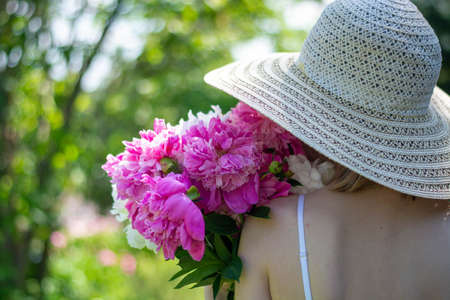 A beautiful girl in a white sundress and a hat is walking along a forest road with peonies in her hands. The concept of a summer holiday.の写真素材