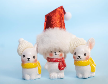 A Christmas card, three ceramic rabbits in knitted hats and a Santa Claus hat stand on a blue background, among Christmas balls and snowflakes, with garland lights. the concept of Christmas.の写真素材