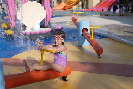A child is playing in a water park. A little girl in a purple swimsuit and swimming goggles, running around the water area of an amusement park during the summer holidays.の写真素材