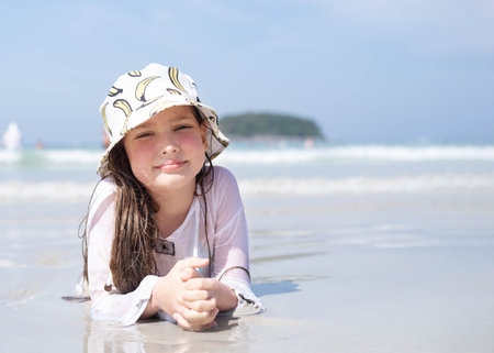 a little girl in a white tunic and hat lies on the beach on a sunny day, against the background of the sea. Summer holidays at the sea.の写真素材