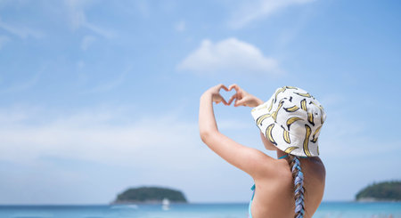 Portrait of a Caucasian child on the background of the sea. A girl stands in a hat and a swimsuit on the beach and shows her heart with her fingers. Have a happy holiday on the sea beach.の写真素材