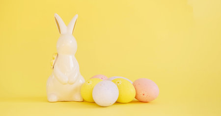 Easter white ceramic rabbit and colored painted eggs isolated on a yellow background. Festive spring...