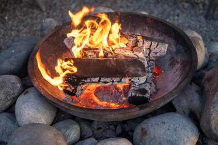 Flames dance around logs in a metal fire pit, surrounded by smooth stones, creating a cozy ambiance...