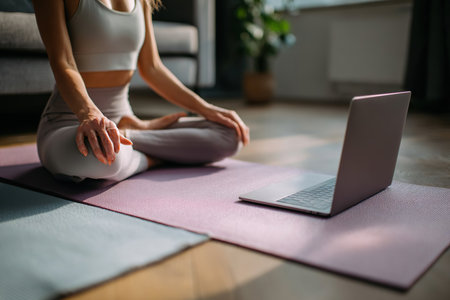 Female yoga practitioner is seated cross-legged on a pink mat in a tranquil indoor environment, with a laptop nearby, enhancing her wellness journey through technologyの素材