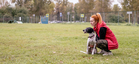 Female dog owner crouches in grassy park beside her pet, demonstrating a bond and connection in a tranquil outdoor setting filled with natureの写真素材