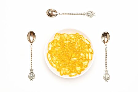 Cod liver oil capsules in a white plate. Omega-3 vitamins in the center in a plate with spoons on the sides on an isolated white background. Natural treatment with antidepressants.の写真素材