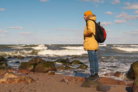 A man in a yellow jacket and hat with a large backpack on the seashore leaned on a large stone and looks into the distance. Traveling in Europe, a world without borders.の写真素材