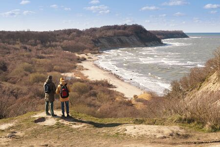 Two men in a yellow jacket and hat with a large backpack, in a green jacket and a dark backpack on a mountainside overlooking the sea or ocean. Traveling in Europe, a world without borders.の写真素材