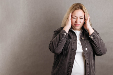 Plus size blonde in a black denim jacket on a gray background holds on to her head in pain.の写真素材