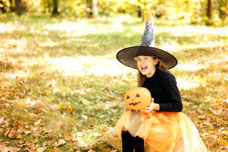 A funny teenage girl in a witch costume and a pumpkin in her hands, laughs sitting in the forest. Kids on Halloween.の写真素材