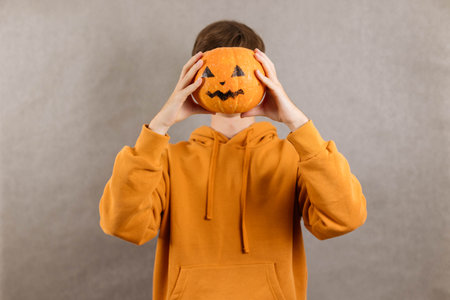 Portrait of a young man on a gray background covering his face with a pumpkin for Halloweenの写真素材