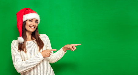 Attractive girl in a Christmas cap points with a finger, the second hand on the belt, green background, copy spaceの写真素材