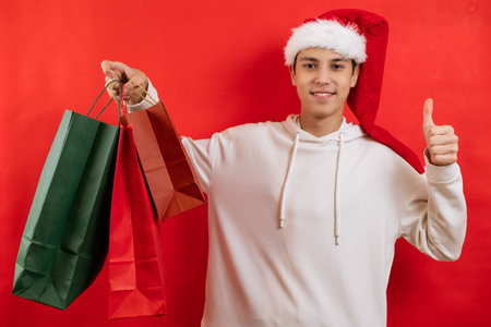 Portrait of a young man in a santa cap and with gifts in his hands smiling on a red backgroundの写真素材