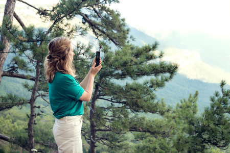 Close-up portrait of a young female tourist in the mountains, takes a photo on a smartphone.の写真素材