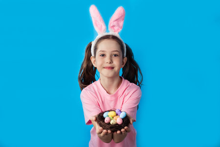 Beautiful little girl in a pink t-shirt with bunny ears holding a nest with easter eggs on a blue background, copy space.の写真素材