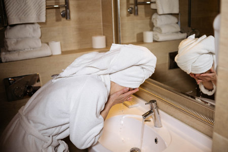 A young beautiful woman washes her face in a stylish bathroom, in a white coat and a towel on her head, skin care treatment conceptの写真素材