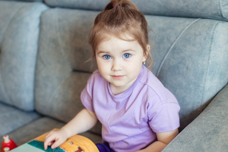 Close-up portrait of cute little girl with blue eyes, sitting on a couch in a room with a book and looking at the cameraの写真素材