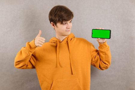 Close-up portrait of a young man on a gray background with a smartphone with a green screen in his...