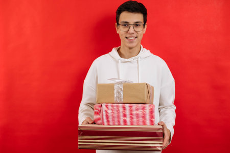 Cute young guy holding a box with a gift on a red background, smiling and looking at the camera.