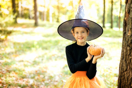 Cute teen girl in hat and witch costume holding pumpkin in autumn forest copy space