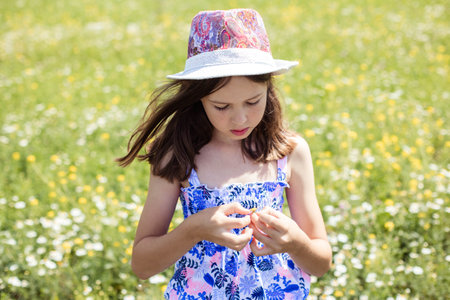 portrait of a teenage girl in a hat on a field background, holding a small flower in her hands and looking downの写真素材
