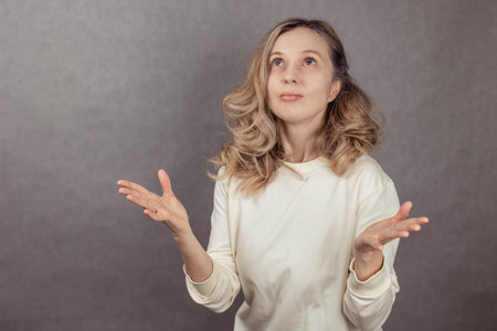 Young woman on gray background looking up in surprise, holding hands to the side, copy space