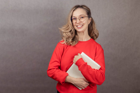 Close-up portrait of young woman in glasses on gray background, holding laptop, smiling and looking...