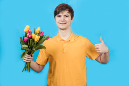 Smiling young man with bouquet of flowers in hands on blue background, looking at cameraの写真素材