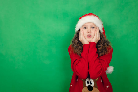 Surprised teenage girl in santa hat on green background holding hands to face, new year, christmas...