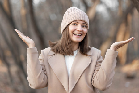 Happy woman in a beige coat and knitted hat, joyfully raising hands in a tranquil outdoor environment, surrounded by trees and nature, expressing delight and warmthの写真素材