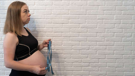 Young pregnant girl in black underwear on a background of a white brick wall. Measuring the growing belly. Copy space.の写真素材