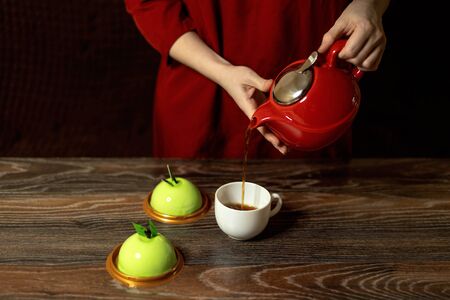 Female hands on the background of a red dress pour tea from a red teapot into a white mug. On the table are cakes.の写真素材