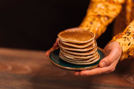 Female hands in a yellow dress hold out a saucer with pancakes with honey. Against the background of...