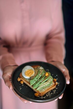 The hands of a girl in a pink dress hold a pink plate with a whole grain bread sandwich, avocado and egg, chickpeas and sesame seeds. Breakfast for proper nutrition.の写真素材