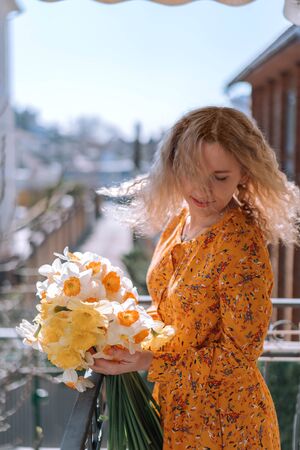 Beautiful young girl on the balcony of the house rejoices at a bouquet of spring flowers, daffodils.の写真素材