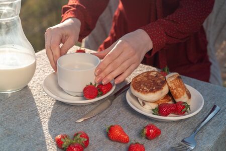 Female hands hold a mug with milk on a table with cheesecakes and strawberries in the fresh air on nature.の写真素材