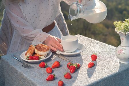 Female hands hold a mug with milk on a table with cheesecakes and strawberries in the fresh air on nature.の写真素材