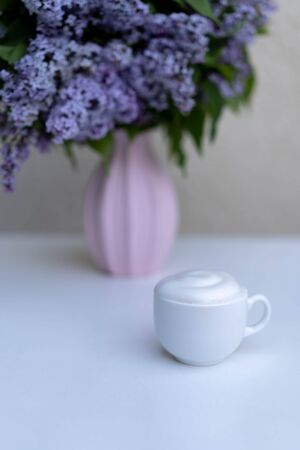 A cup of morning drink with whipped cream on a background of a bouquet of lilacs in a pink vase.の写真素材