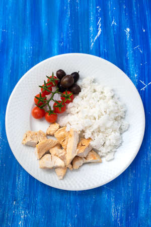chicken breast with boiled rice and tomato on white plate on blue woodの写真素材