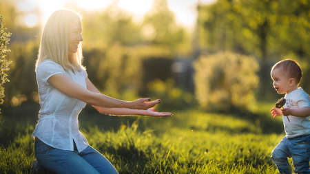 The kid runs through the grass into the arms of his mother in the park at sunsetの写真素材