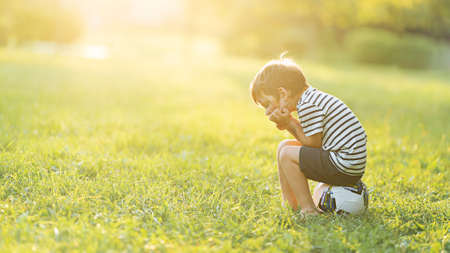 A little boy sits on a soccer ball and is sad on a grassy field with a sun glare.の写真素材