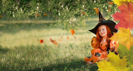 Little girl in a witch costume is smiling while sitting in the park on the street with a frame of autumn leaves with copy spaceの写真素材